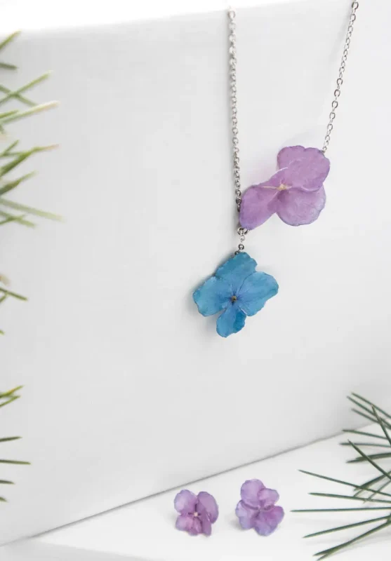 نیم ست گل طبیعی رزینی طنین با گل‌های هورتانسیا یاسی و آبی روی زنجیر استیل، همراه با گوشواره‌های گل هورتانسیا رزینی در کنار شاخه‌های سبز تزیینی